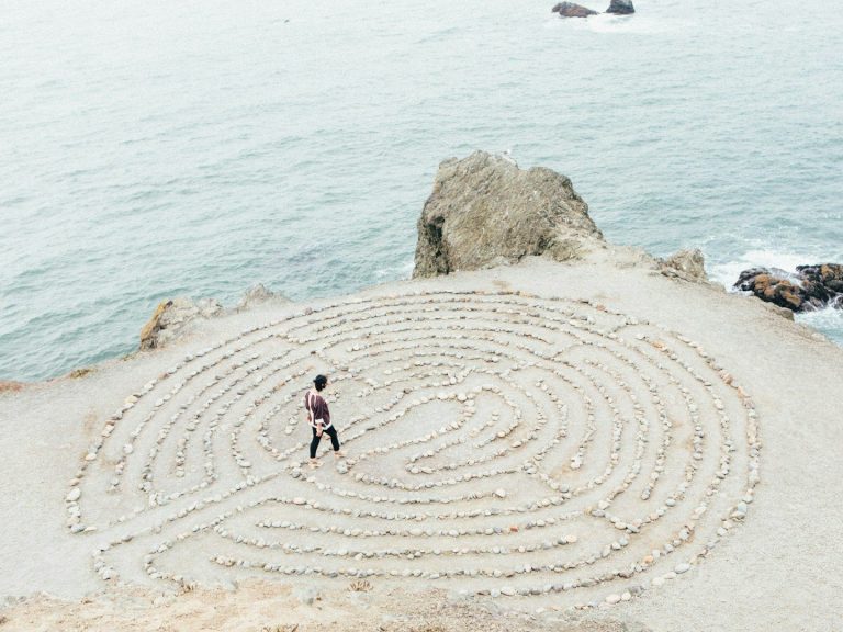 Een persoon wandelt door een zandlabyrint aan de kust. Coach en therapeut.